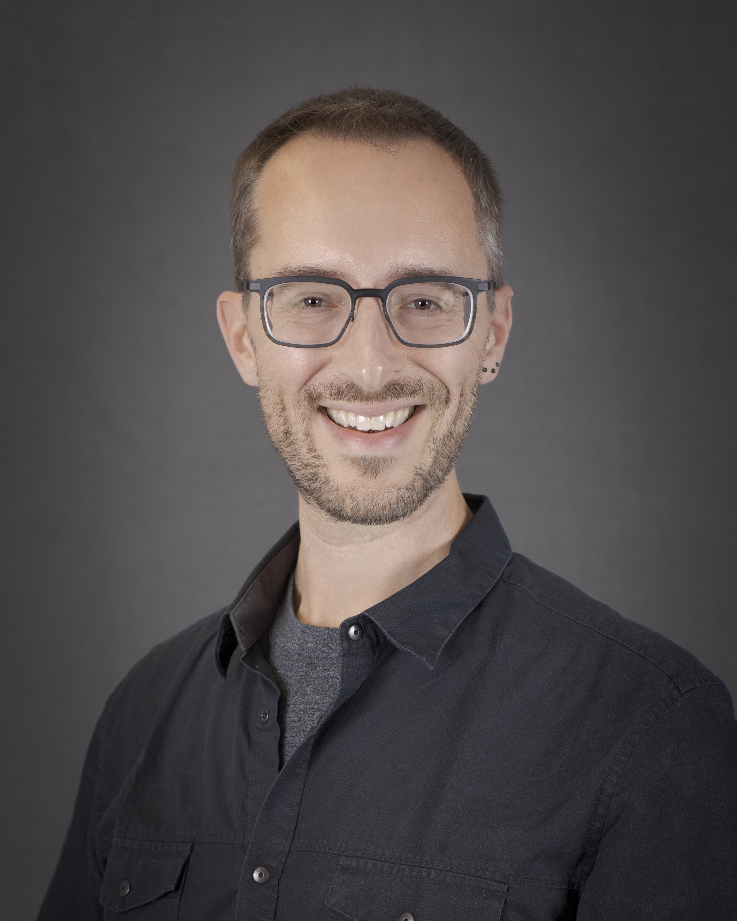 A portrait of a person with short black hair wearing a black collared shirt and glasses