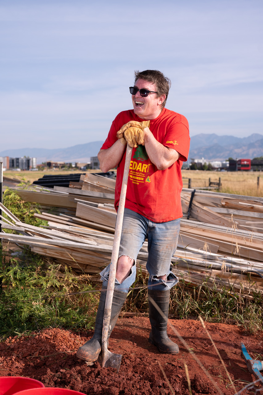 A person with short brown hair wearing a red shirt and ripped blue jeans with one foot on a shovel