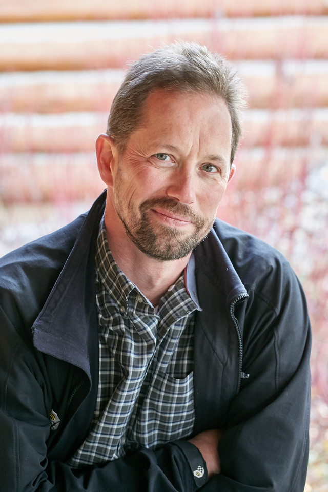 A portrait of a person with dark colored light facial hair and a black collared coat